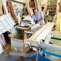 Man working in a cluttered woodworking shop with a table saw and stacked lumber