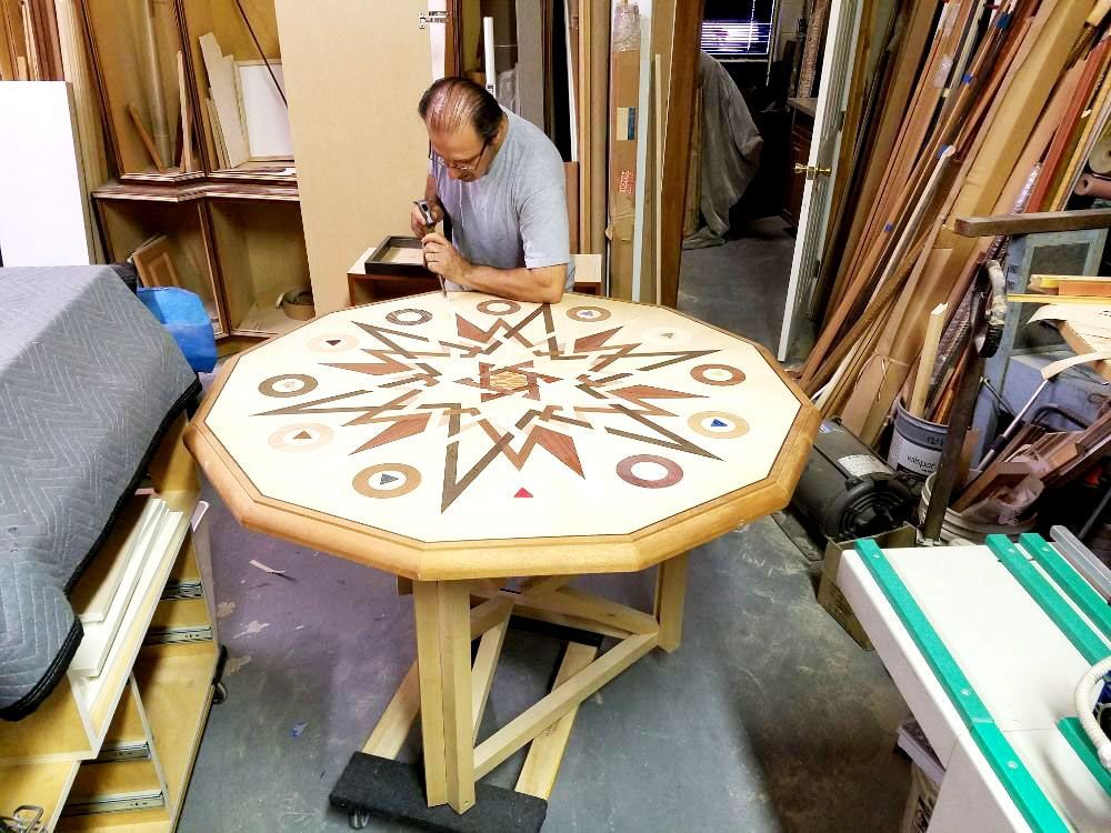 Man working on an octagonal wooden table with a geometric inlay in a workshop, surrounded by lumber.