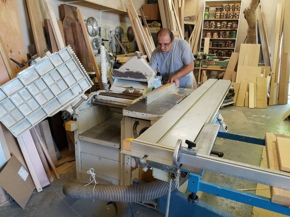 Man working at a woodworking table saw in a cluttered workshop, with lumber and tools around him.