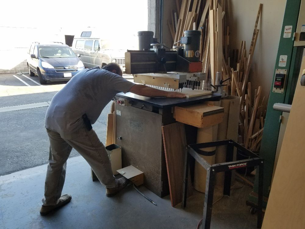 Person leaning over workbench in a workshop, operating a power tool beside stacked wood and tools.