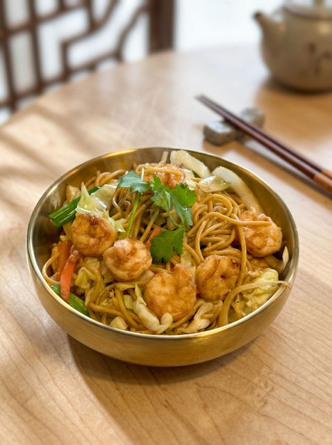 Bowl of fried noodles with shrimp and vegetables on a wooden table, with chopsticks beside it