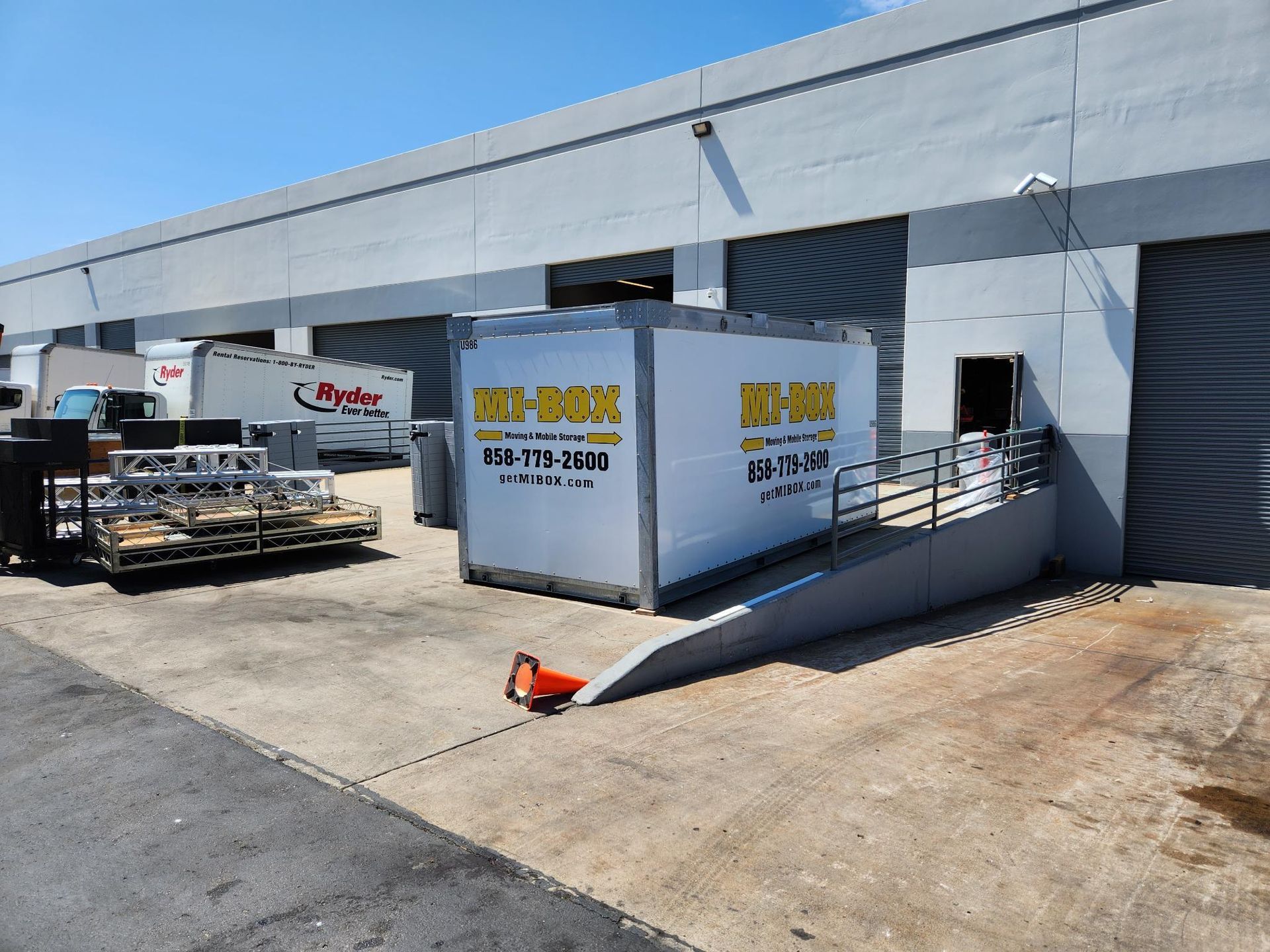 Industrial building with a large sign, white trailer, and fenced utility area beside a paved driveway.