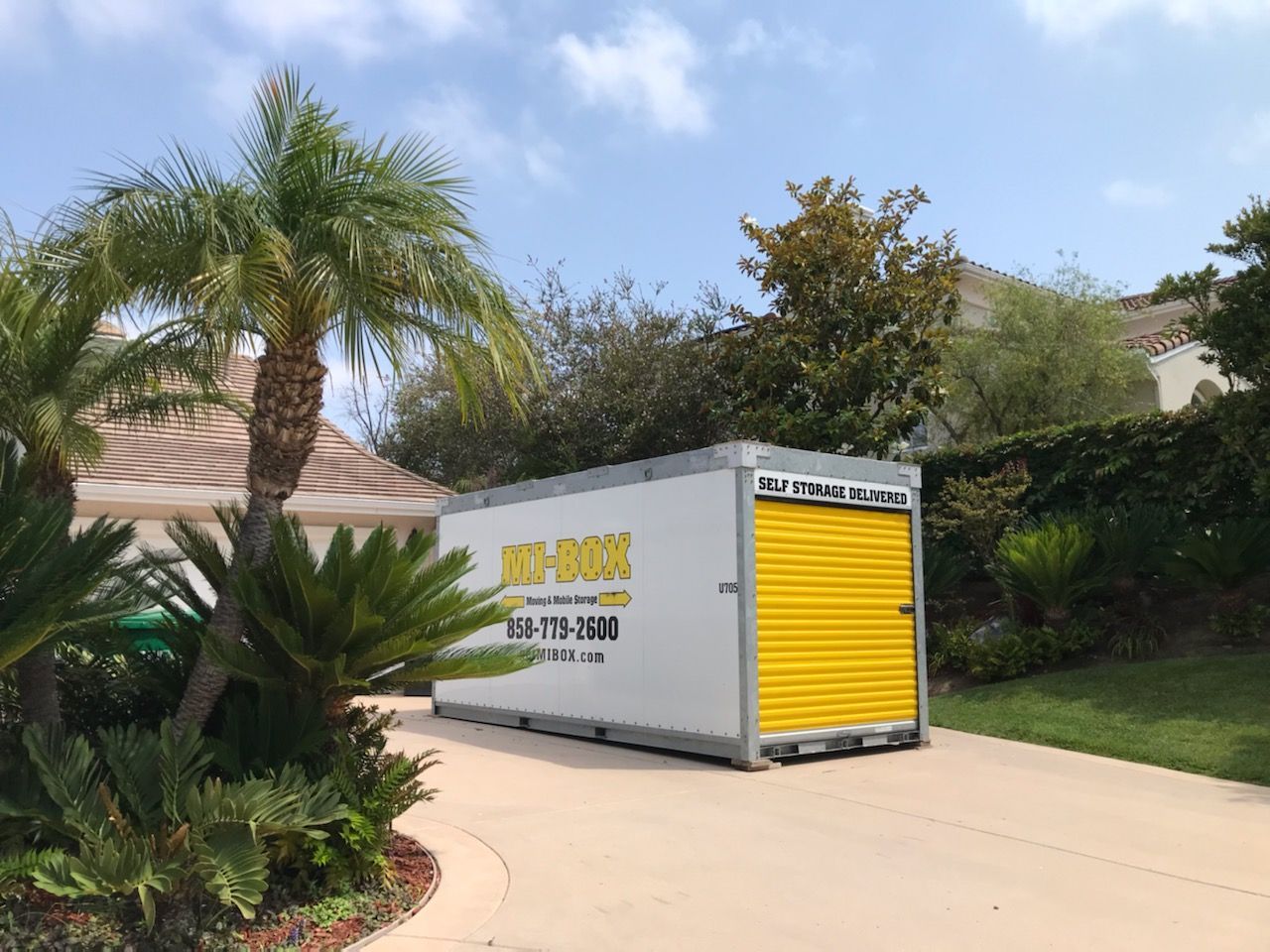 A white container with a yellow door is parked in a driveway next to a palm tree.
