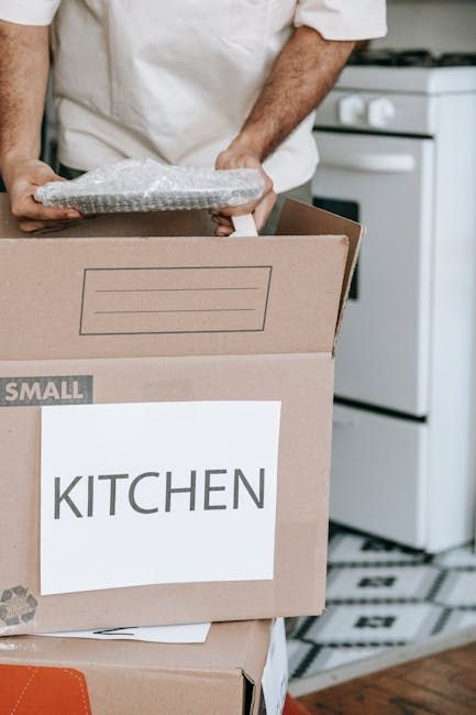 Person carrying a box labeled “KITCHEN” during a move in a kitchen.