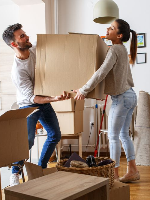 Two people carrying a large cardboard box in a bright moving room, with moving supplies nearby