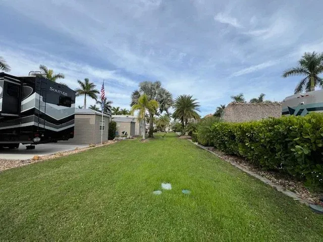 RV park with green lawn, palm trees, and RVs under a partly cloudy sky.