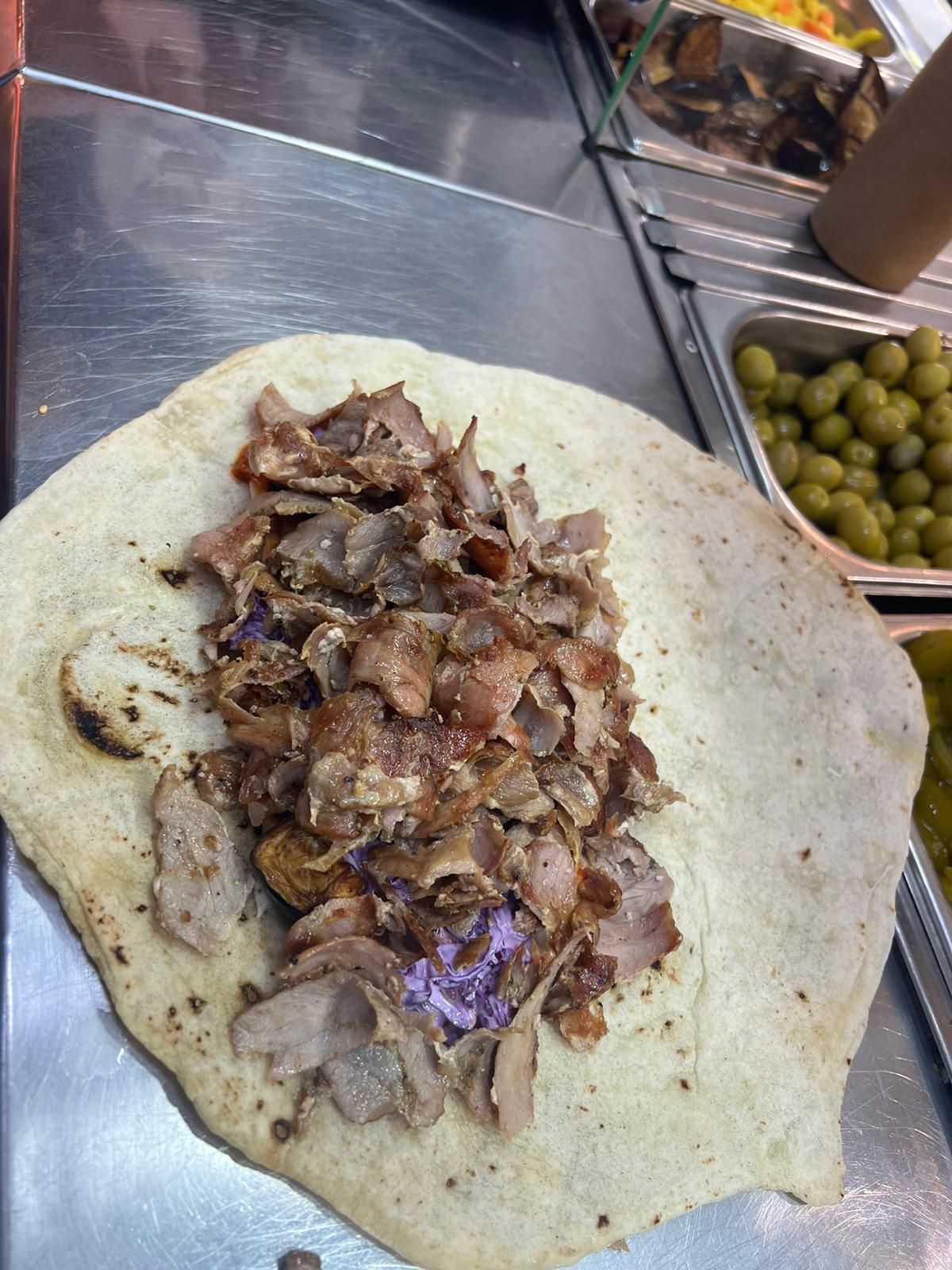 לחם שטוח עם בשר צלוי קצוץ על משטח מתכת, עם זיתים ירוקים ברקע