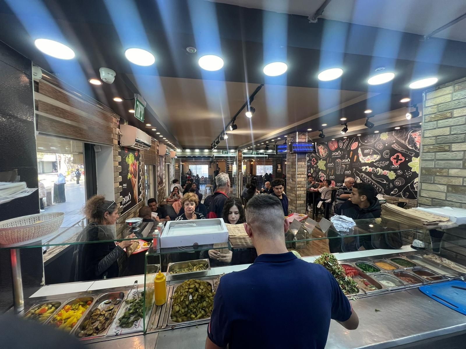 דלפק מעדנייה עמוס בלקוחות, מגשי תצוגה מזכוכית ותאורה בהירה מעל שוק אוכל.