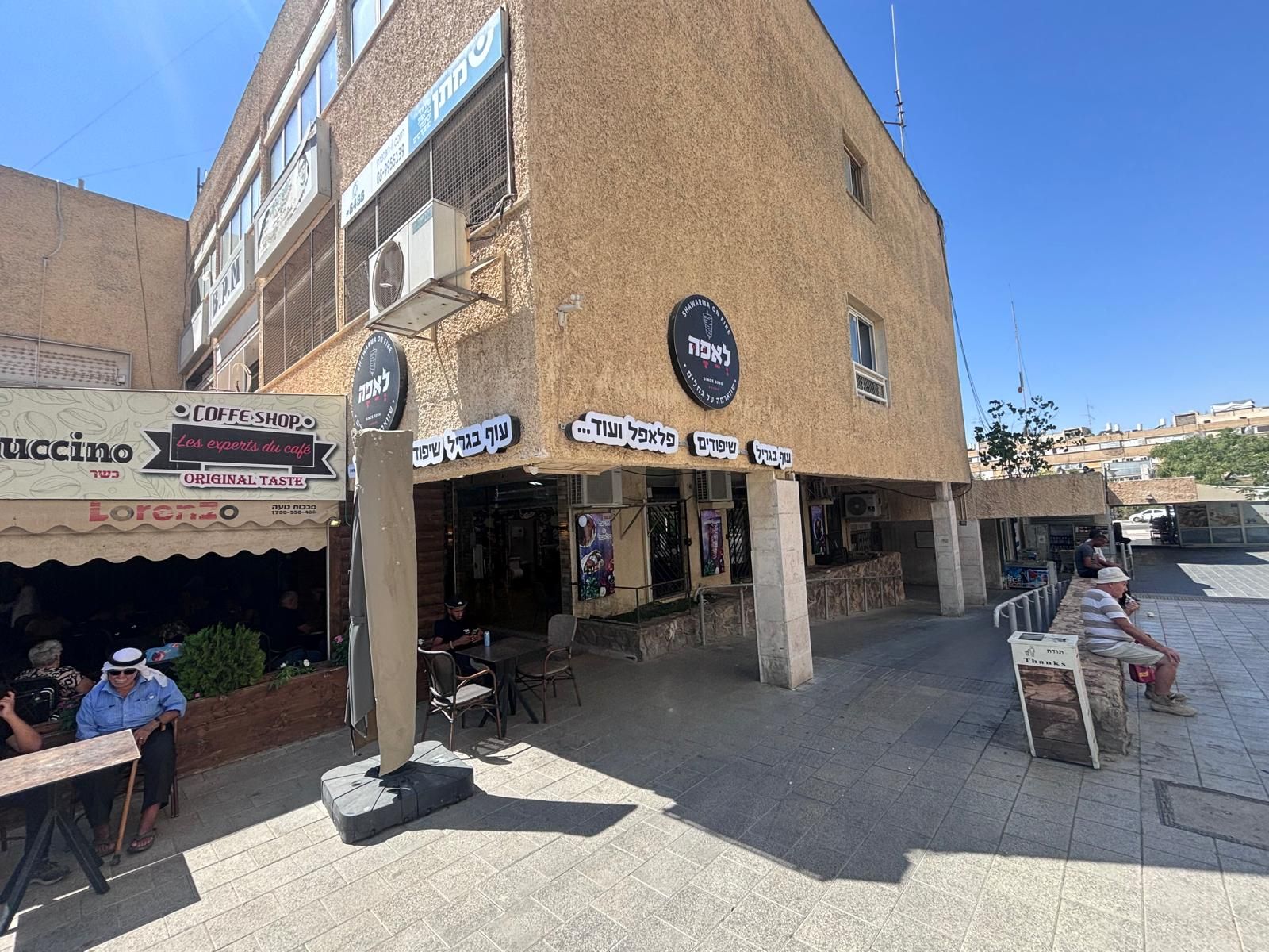 מרפסת בית קפה חיצונית ליד בניין טיח חום בהיר עם אנשים יושבים תחת שמשיות וצל.