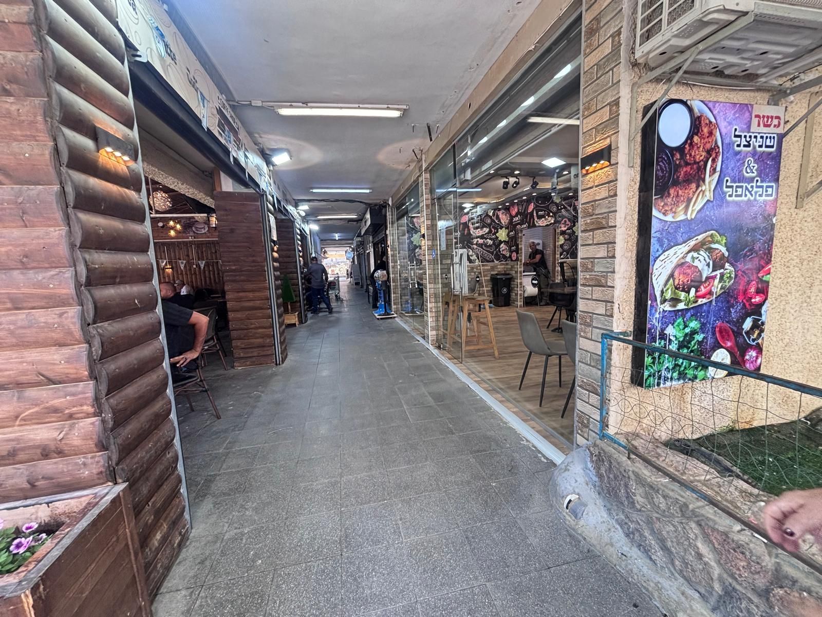 מסדרון שוק מקורה וצר עם דוכני חנויות וסחורות תלויות תחת אורות פלורסנט.