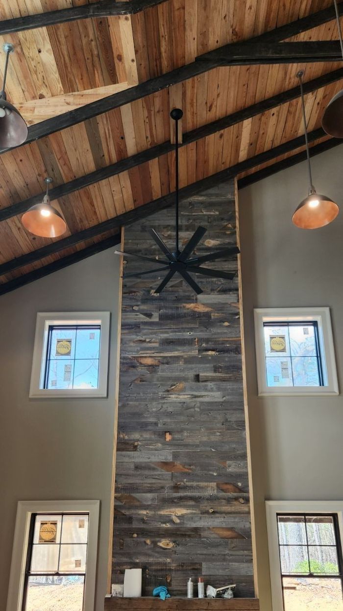 Rustic interior with a wooden ceiling, stone chimney, and windows. A ceiling fan and pendant lights hang down.