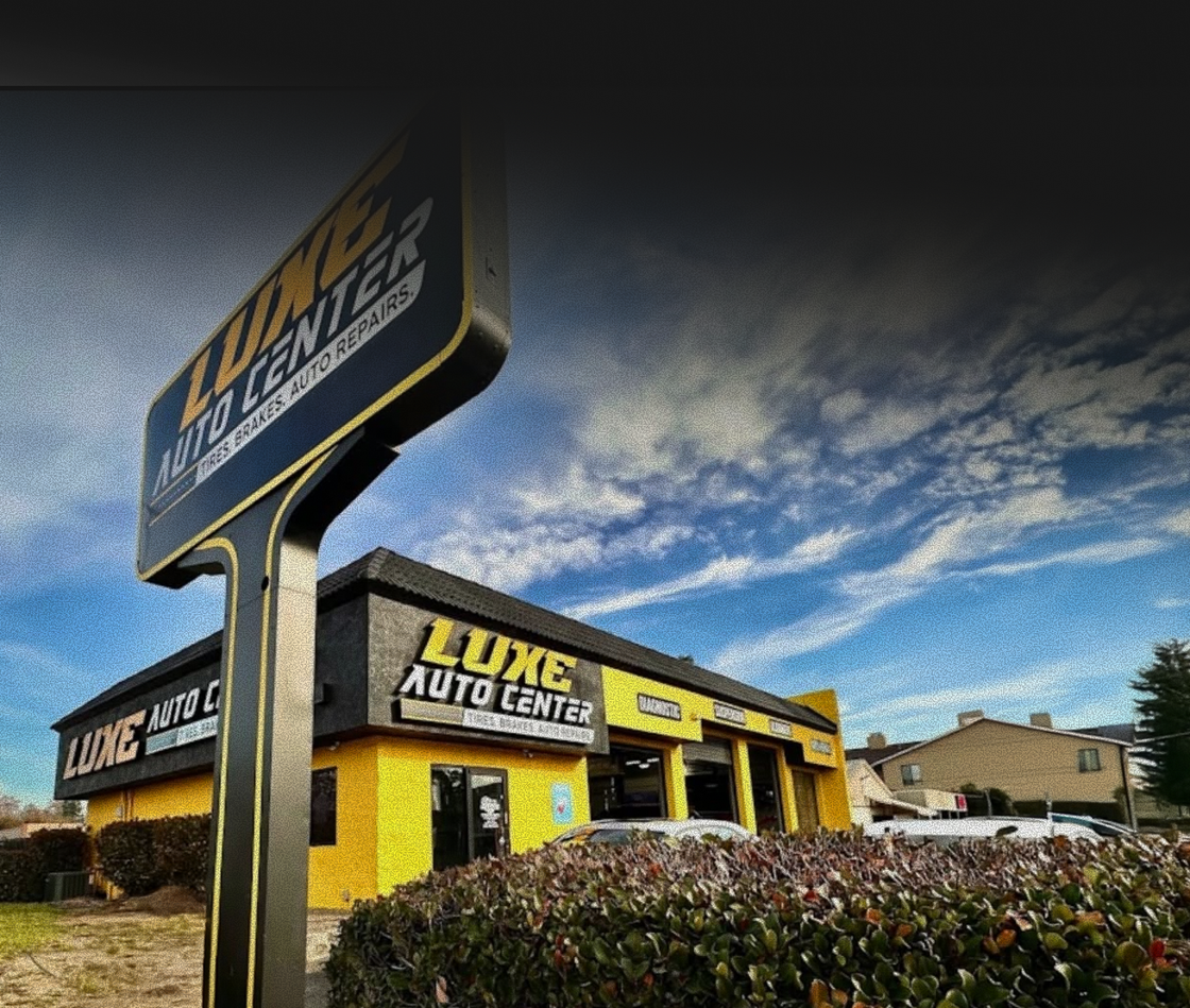 Luxe Auto Center storefront with yellow and black signage behind a chain-link fence and shrubs