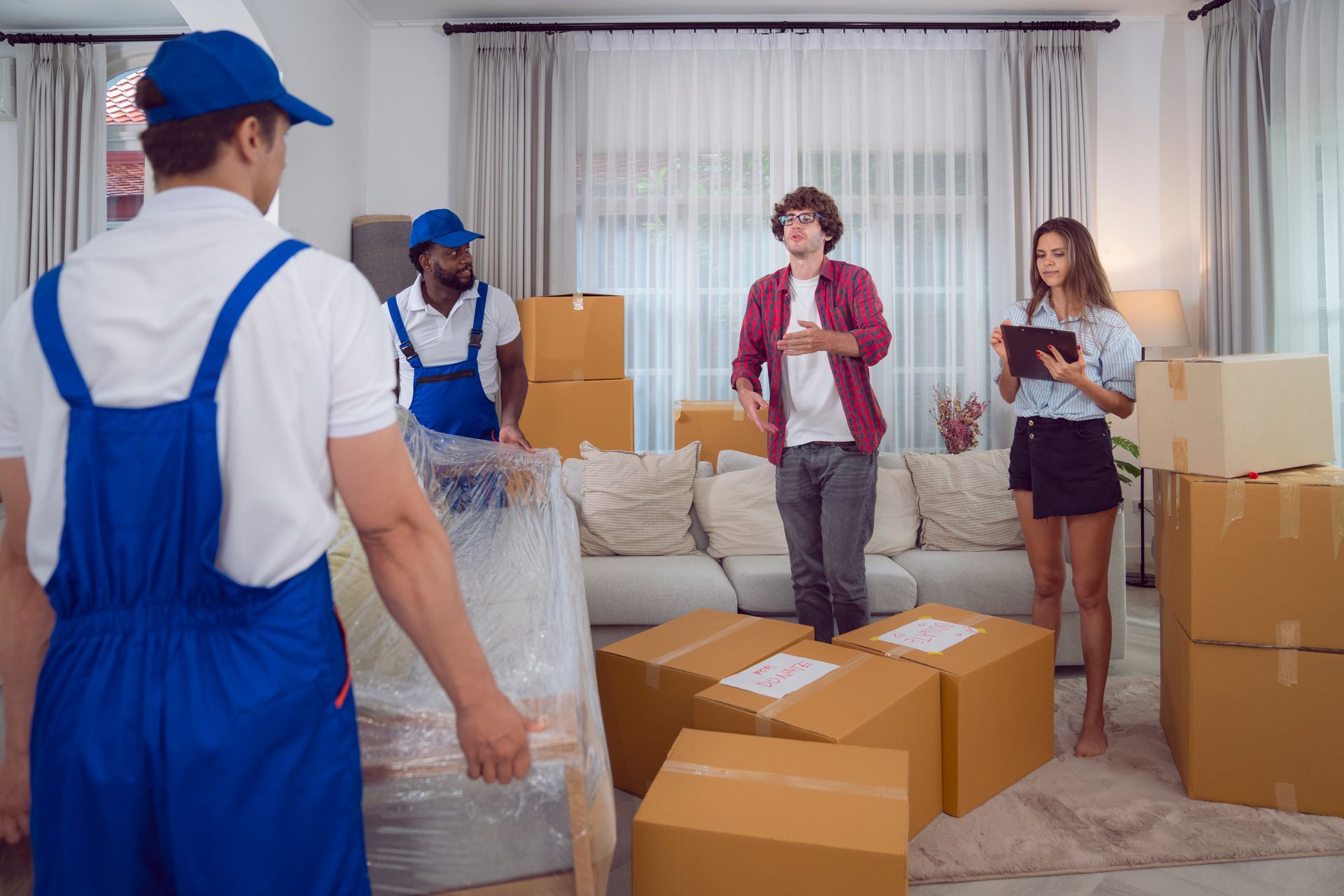 Movers loading boxes in a living room as a couple observes; wrapped furniture, packed boxes.