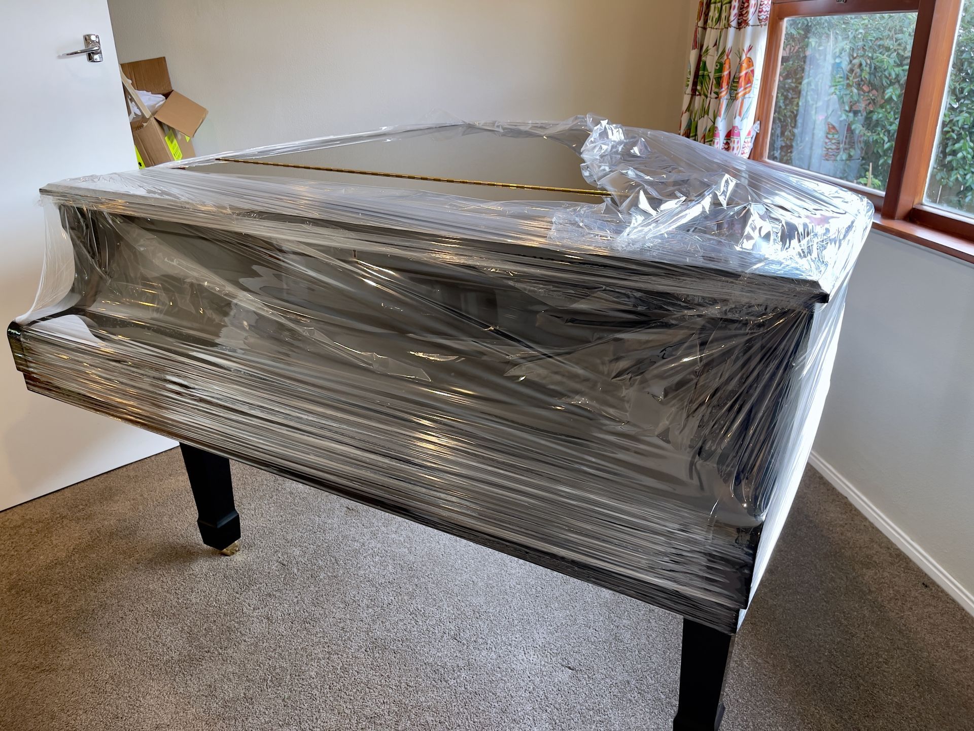 A grand piano wrapped in plastic, in a room with a window and a door.