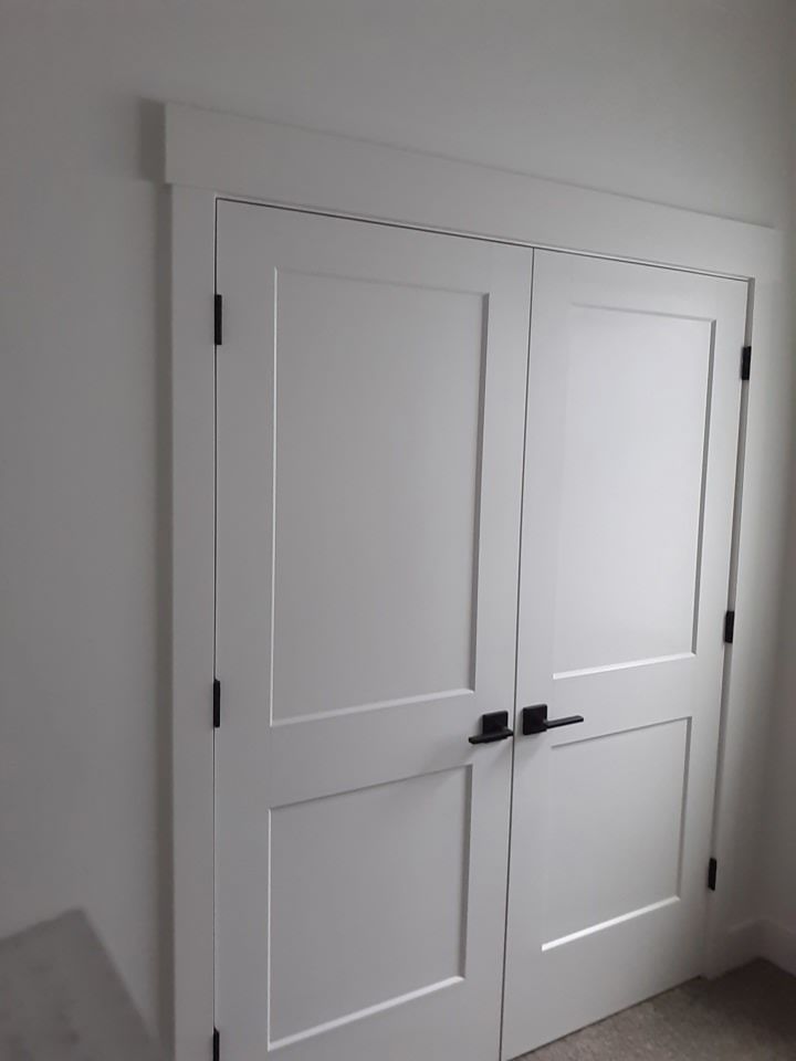 White double doors with black hardware, framed by white molding, against a white wall.