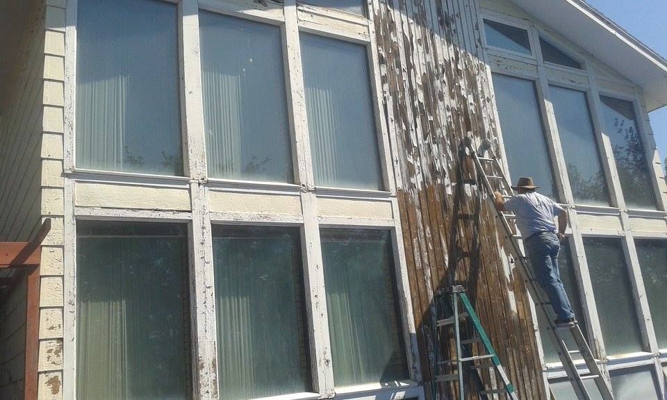 Man on ladder removing weathered wood siding from a house with large windows.
