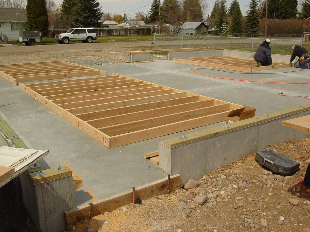 Construction site: wooden floor framing on concrete foundation, two workers, sunny outdoors.