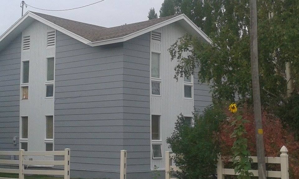 Two-story building with gray and white siding, tall windows, and a brown roof, next to a tree and white fence.