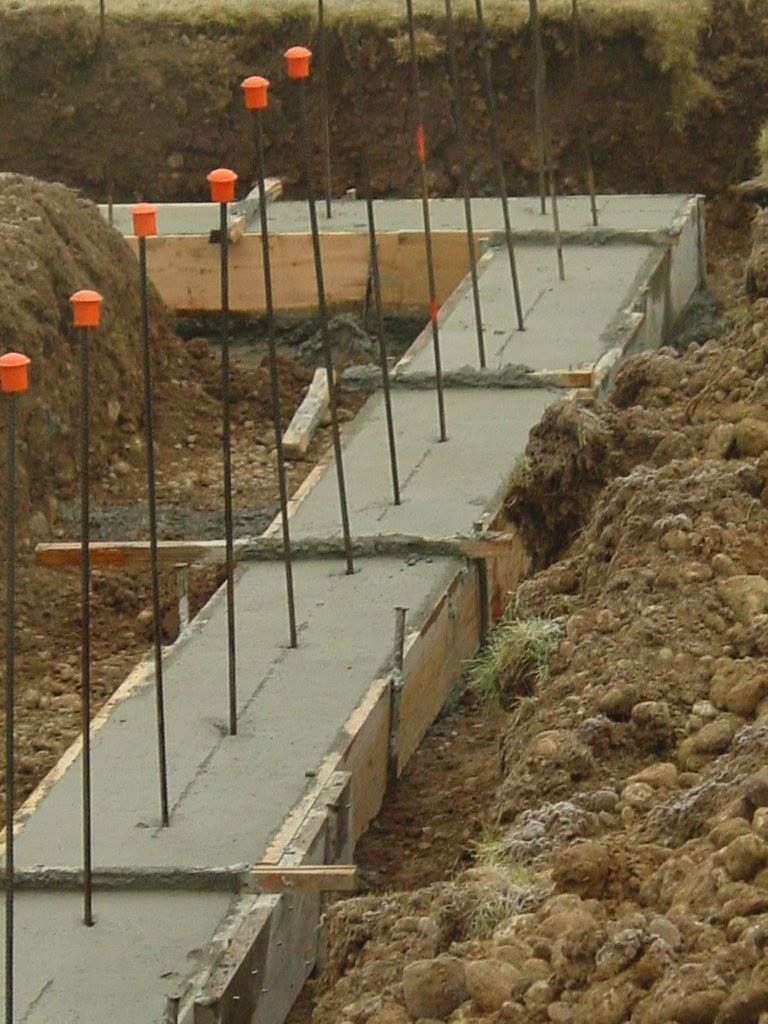 Concrete foundation forms with rebar in an excavated area. Wooden formwork holds fresh concrete.