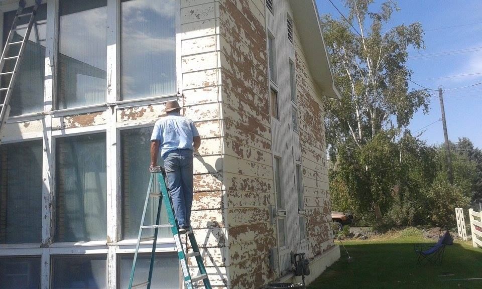 Person on ladder scraping peeling paint off a white house with large windows. Sunny day, green grass.