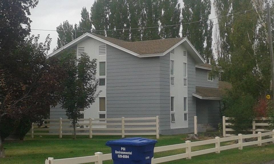 Gray two-story house with white trim and a brown roof, behind a white fence and a blue trash can, with tall trees in the background.