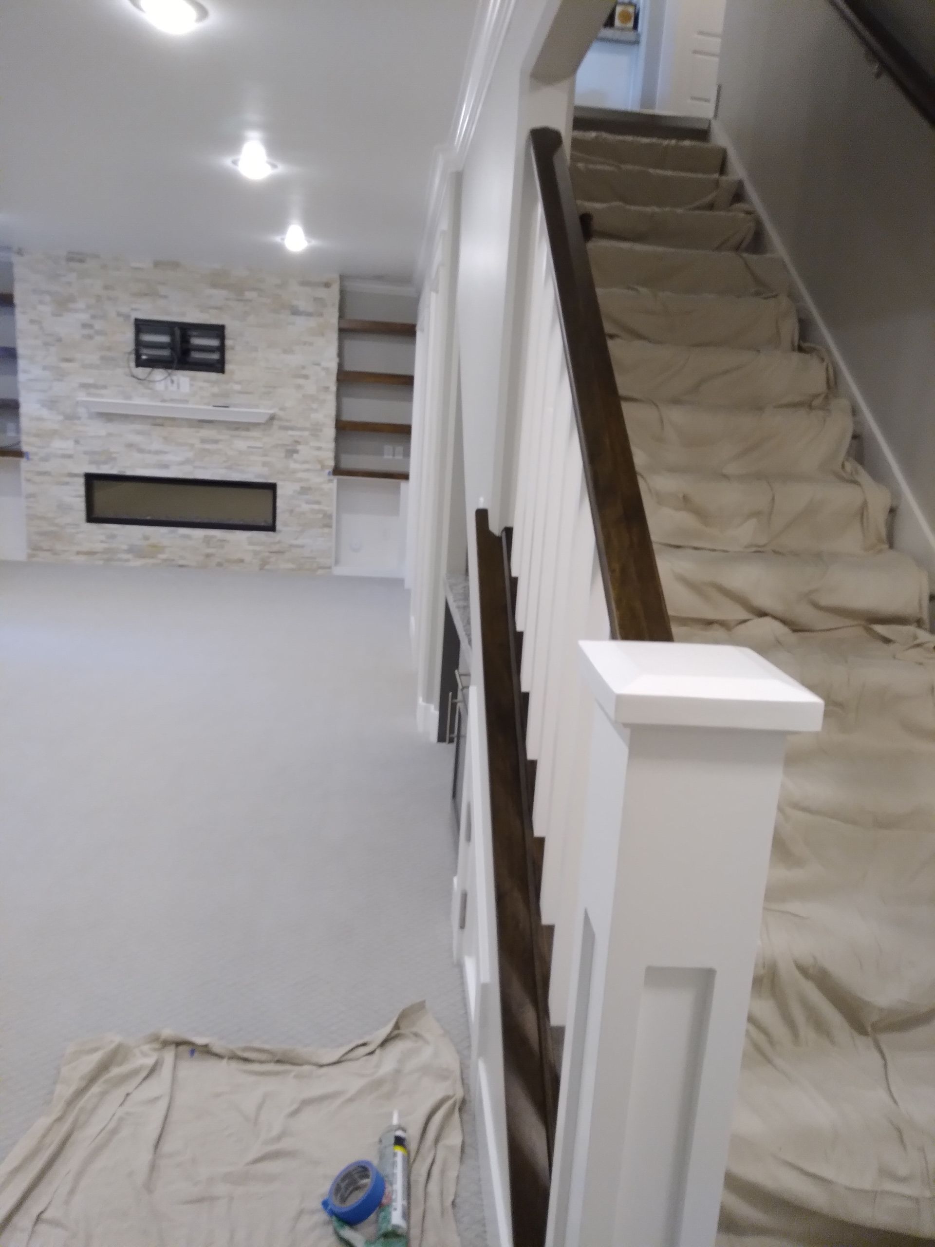 Staircase with brown handrail and white balusters. White walls, grey carpet. Paint supplies on the floor.