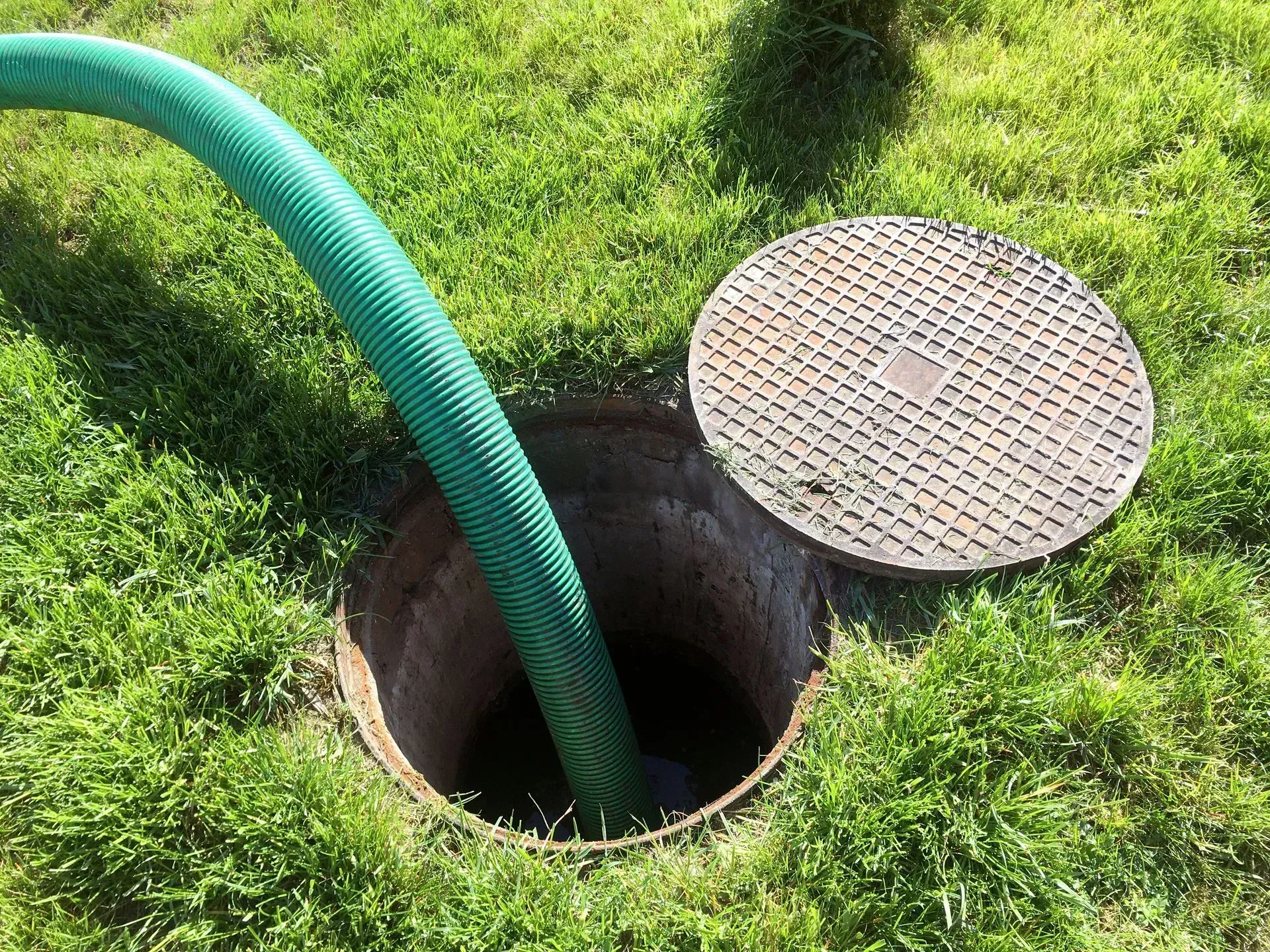 Green hose in an open septic tank with lid on grass.
