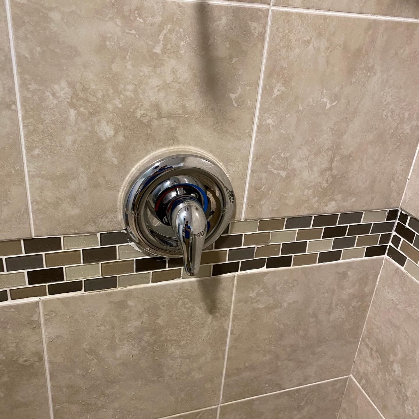 Chrome shower control knob on beige tiled bathroom wall with a decorative mosaic border