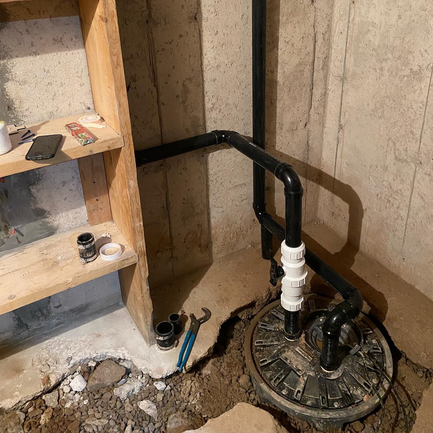 Basement plumbing with black pipes and a round sump pump pit beside unfinished wooden shelving.