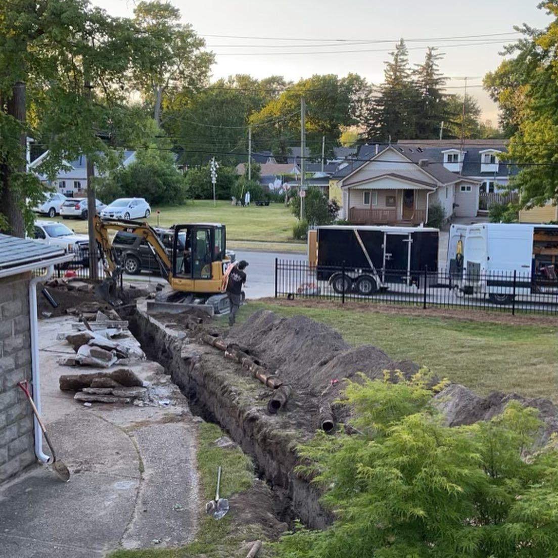 Backhoe digging a long trench beside a house in a suburban yard at dusk