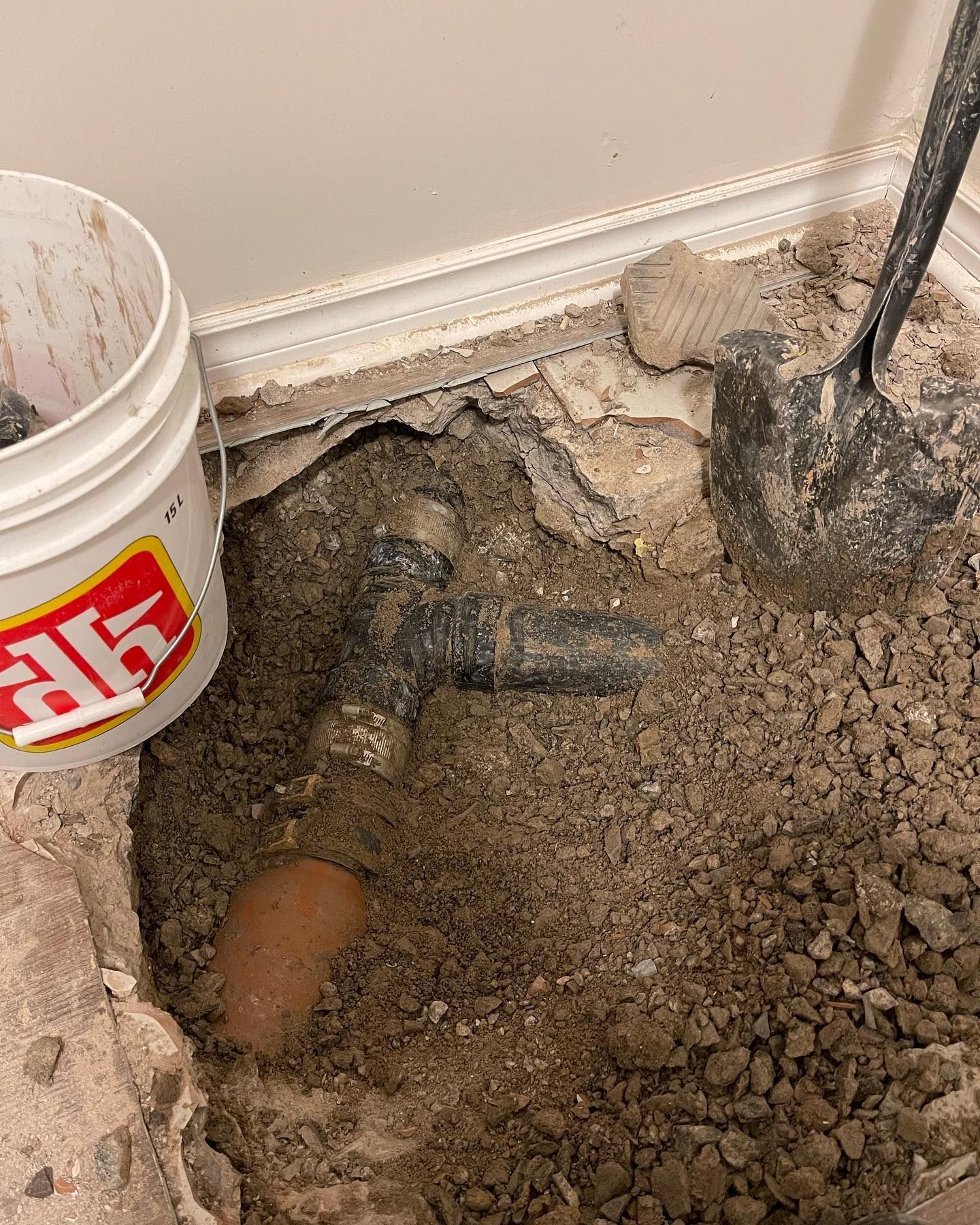 Broken floor exposing a pipe and rough concrete beside a white bucket of mortar or plaster