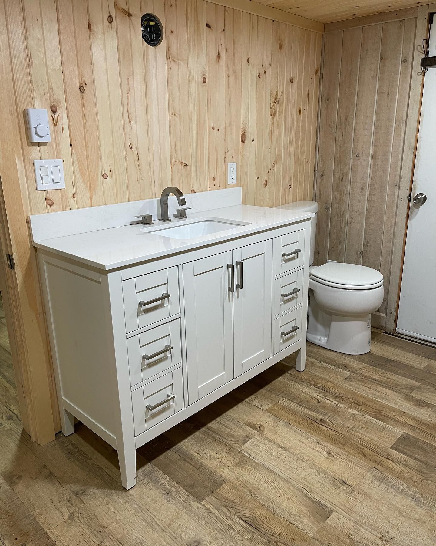 Small bathroom with white vanity and sink, toilet, wood-paneled walls, and wood-look flooring.