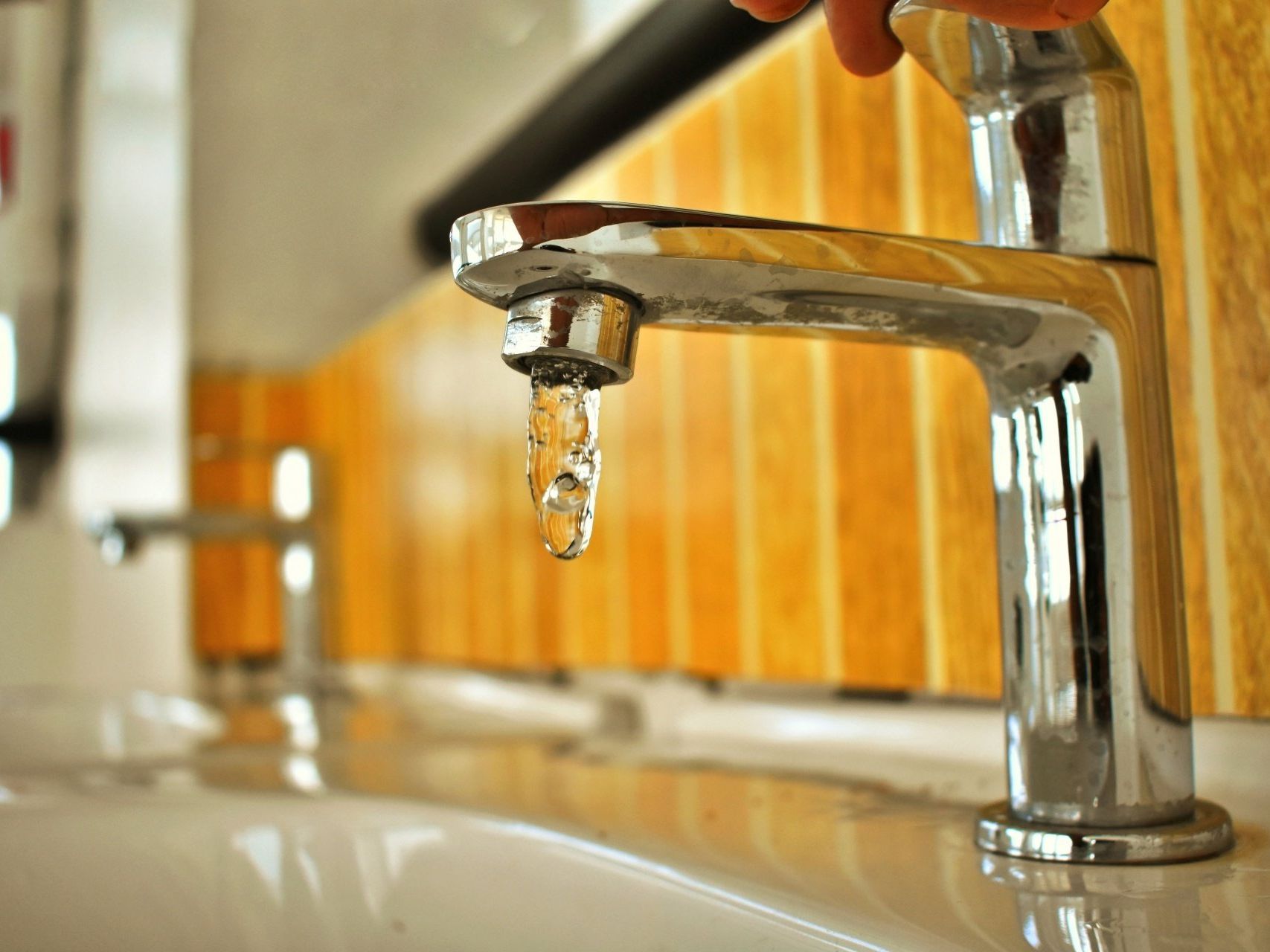 Hand turning off a chrome faucet with a dripping drop in a tiled sink area