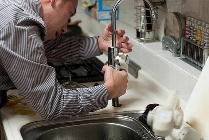 Plumber repairing a kitchen faucet at a sink, with tools and cleaning supplies nearby
