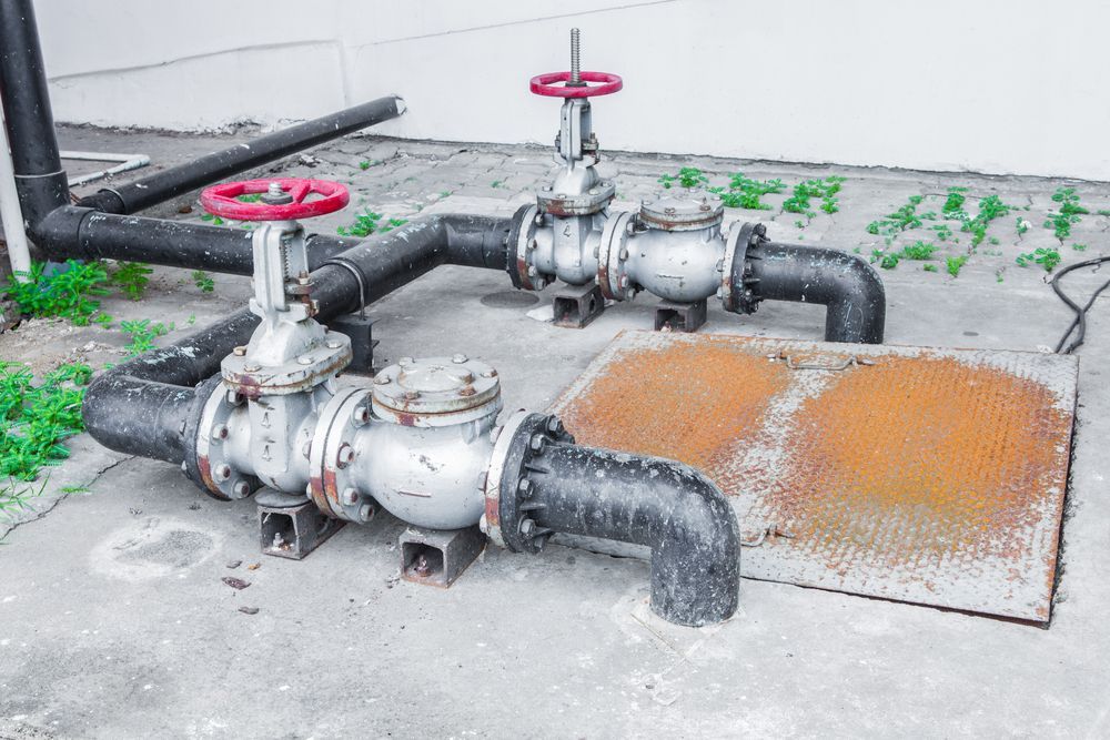 Two red-valve industrial pipes on concrete beside a rust-stained manhole cover