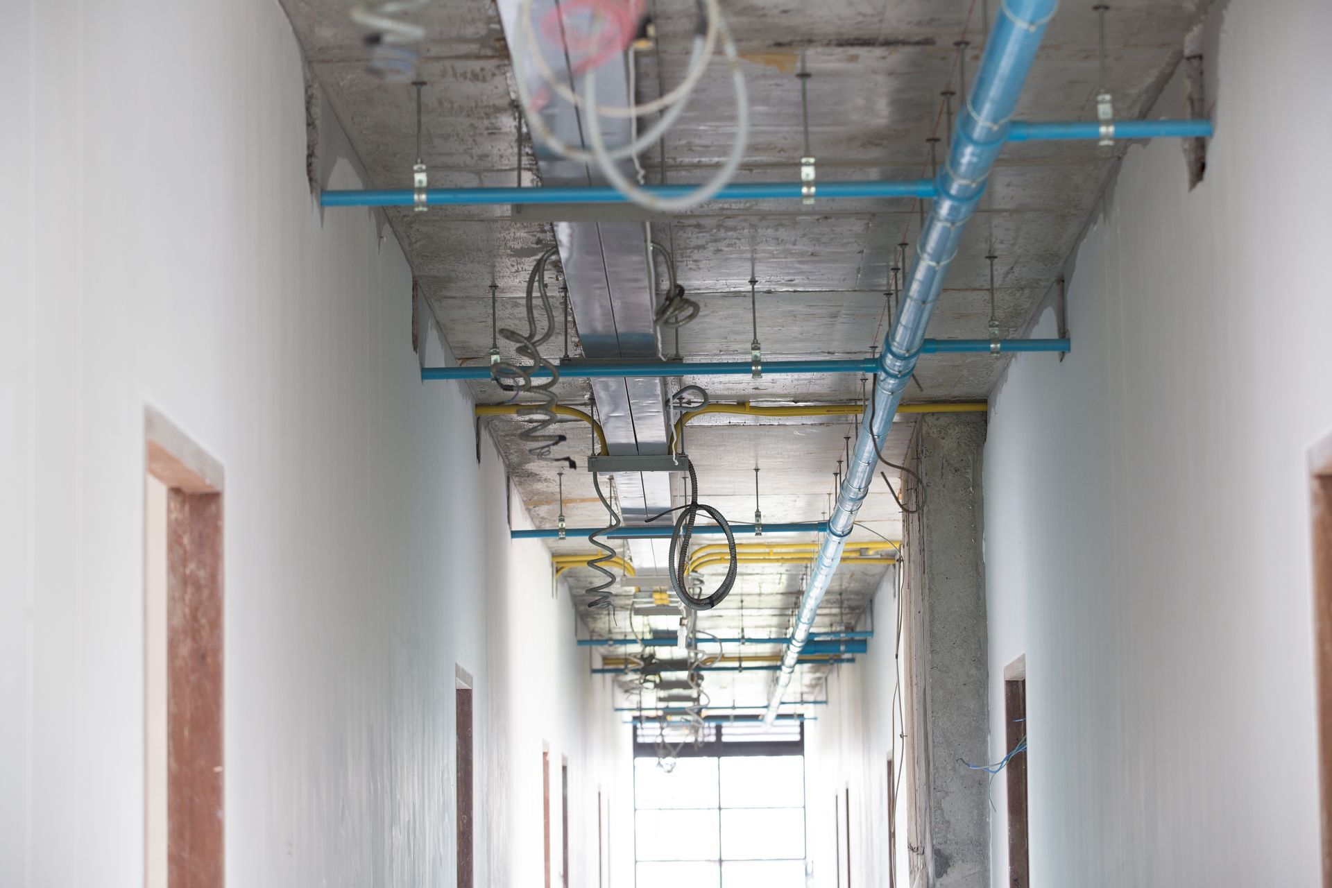 Partially finished hallway with exposed ceiling pipes and wiring, white walls, and doorways on both sides