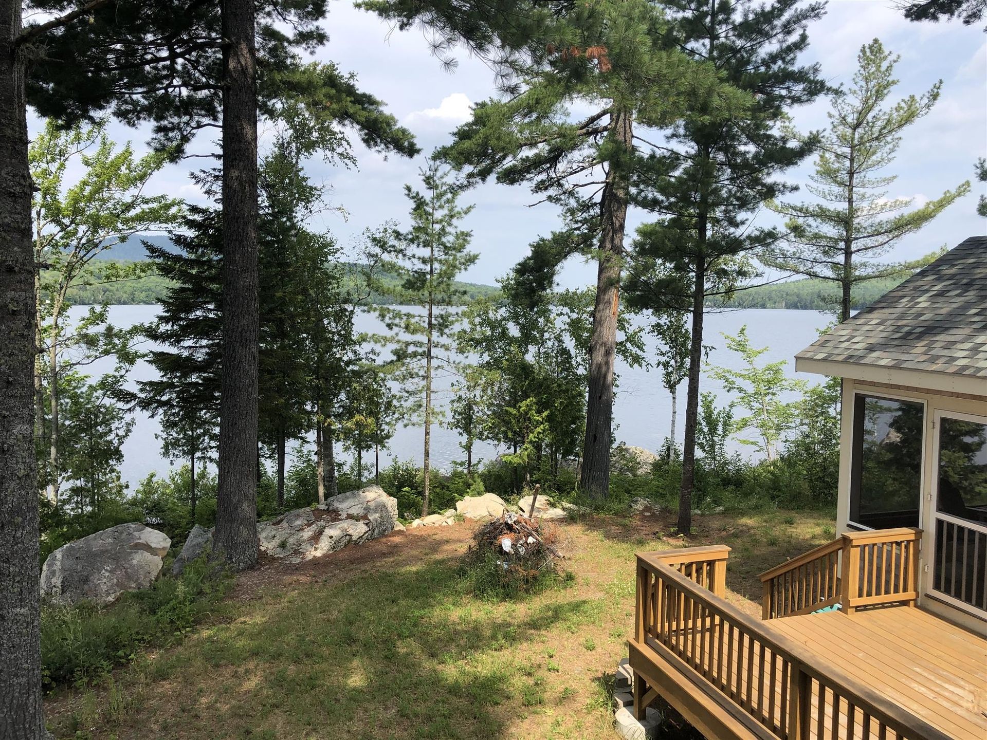 Wooden lakeside deck beside tall pine trees, with a fire pit and water visible through the trees.