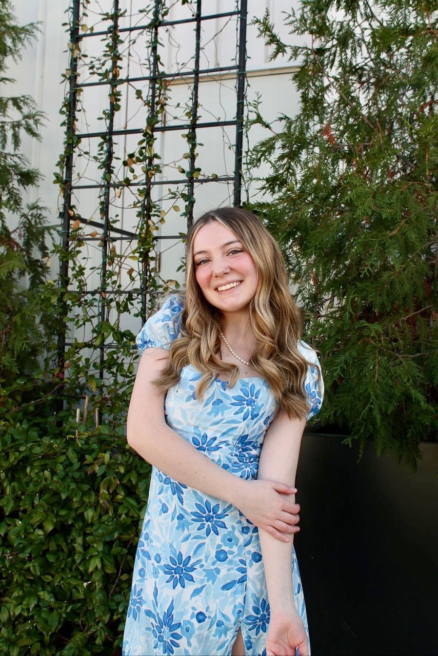 Smiling person in a blue floral dress standing outdoors by a vine-covered wall