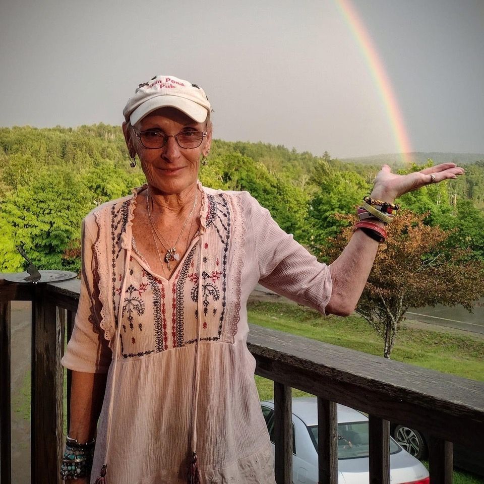 Person on a balcony holding up a hand toward a rainbow over green trees
