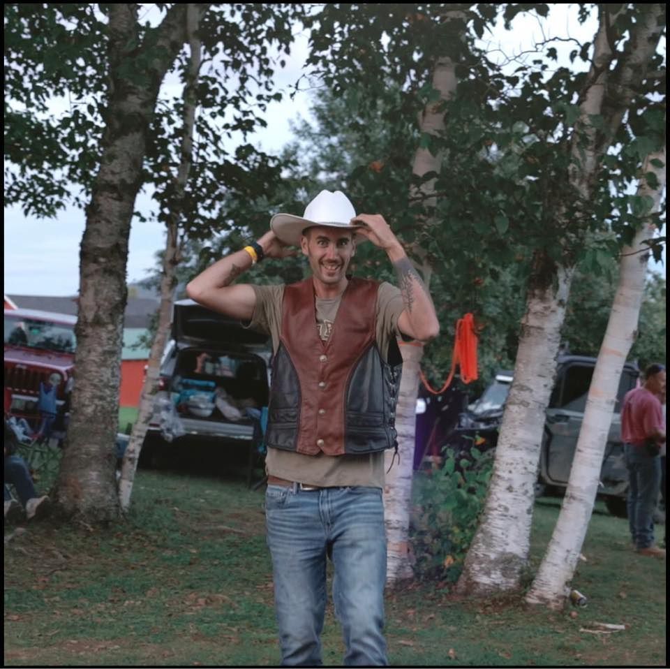 Man in a white cowboy hat stands between trees, adjusting his hat in an outdoor grassy area.