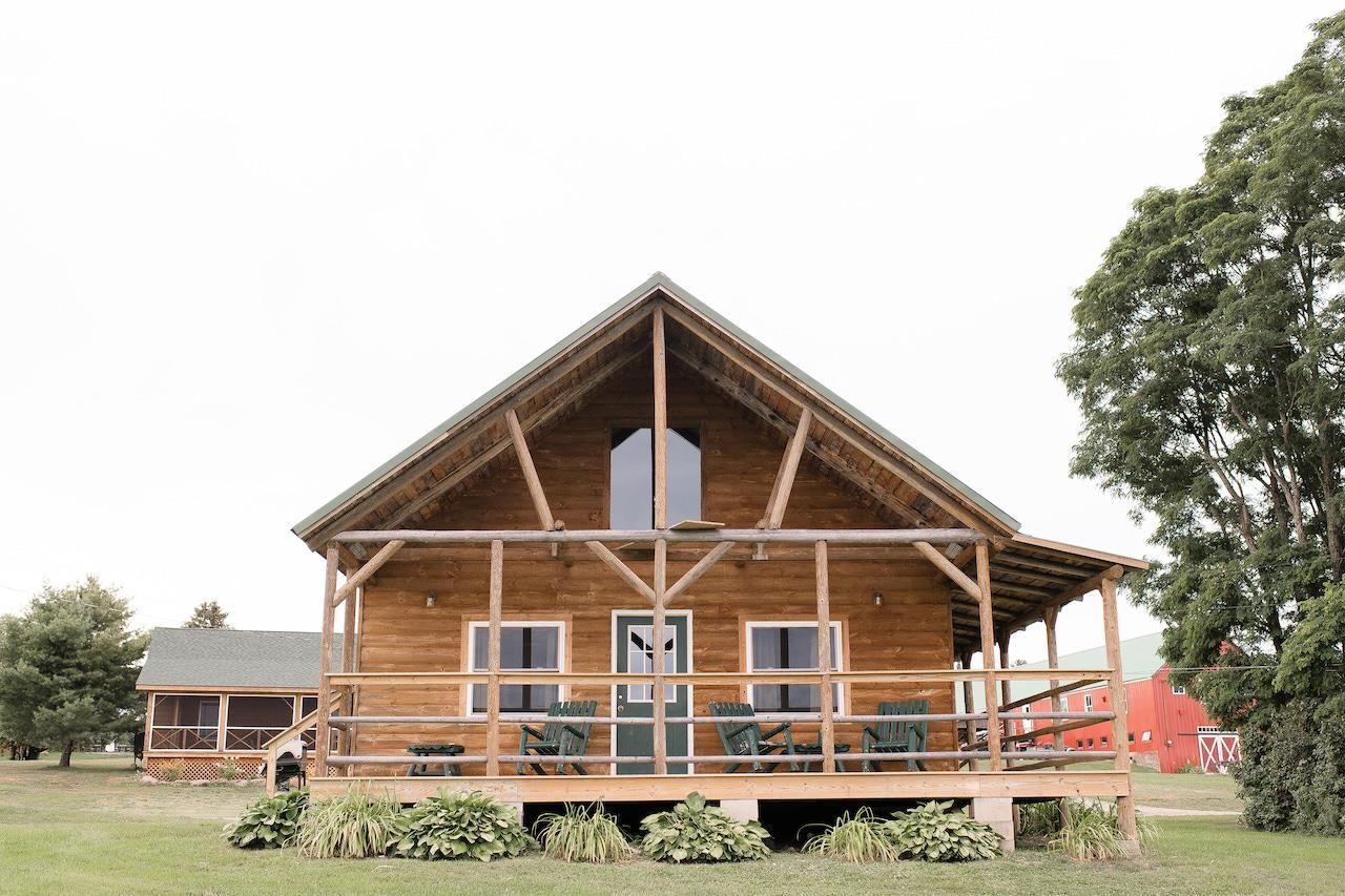 Wooden cabin with a front porch and steep roof, set in a grassy yard with trees nearby.