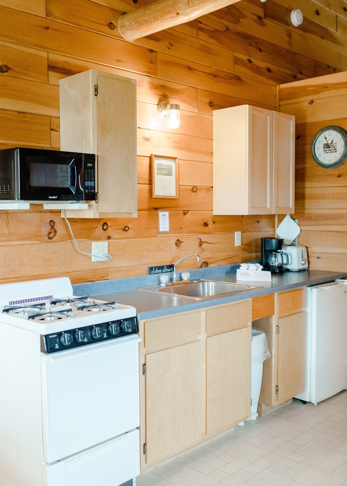 Small rustic kitchen with wood-paneled walls, white stove, microwave, sink, cabinets, and refrigerator.