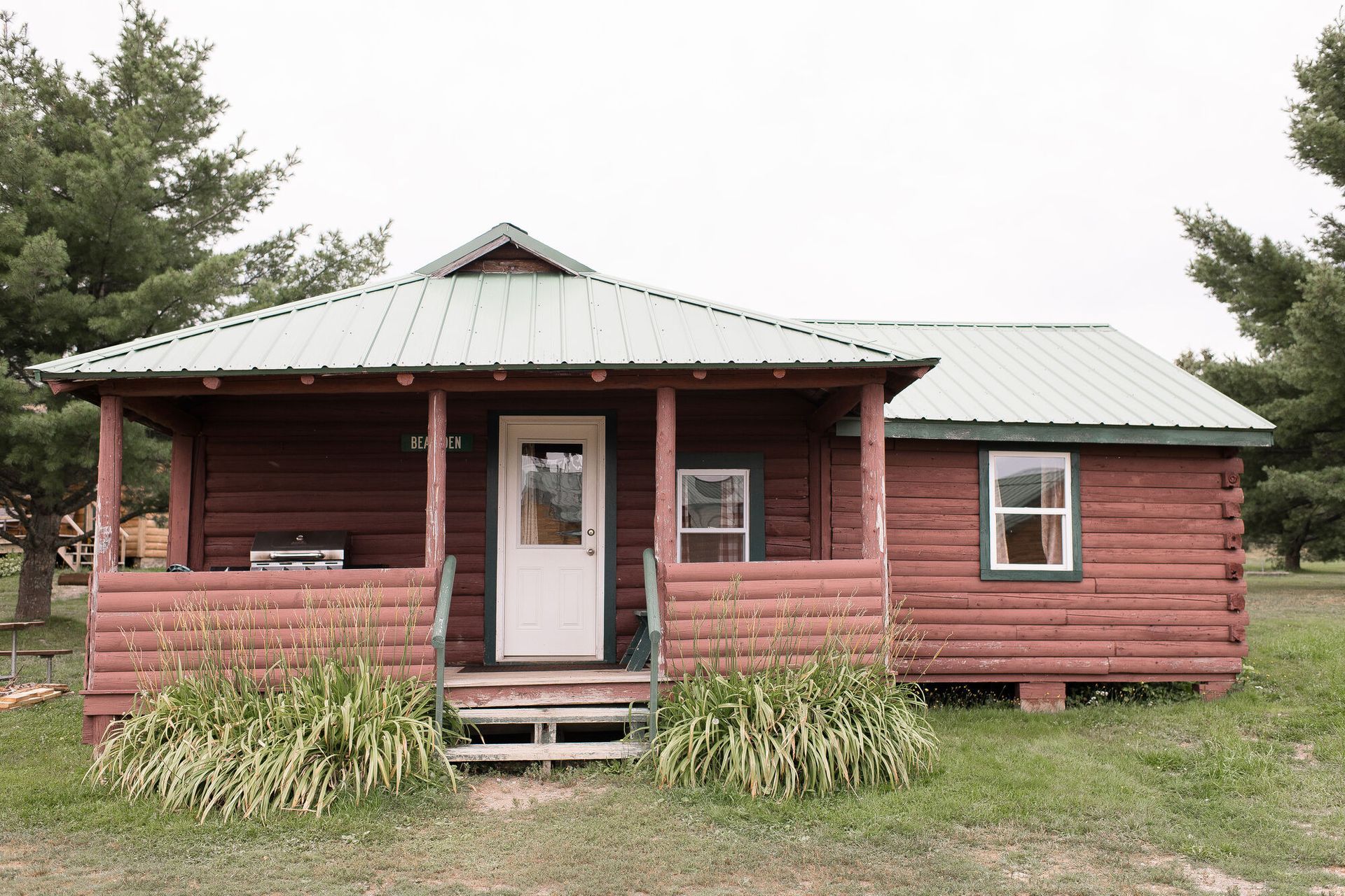 Small red log cabin with a green metal roof, front porch, and grass yard