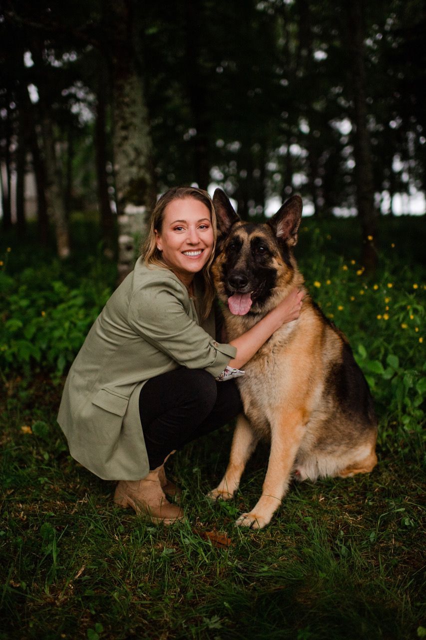 Woman crouches beside a large German shepherd in a grassy, tree-lined park.