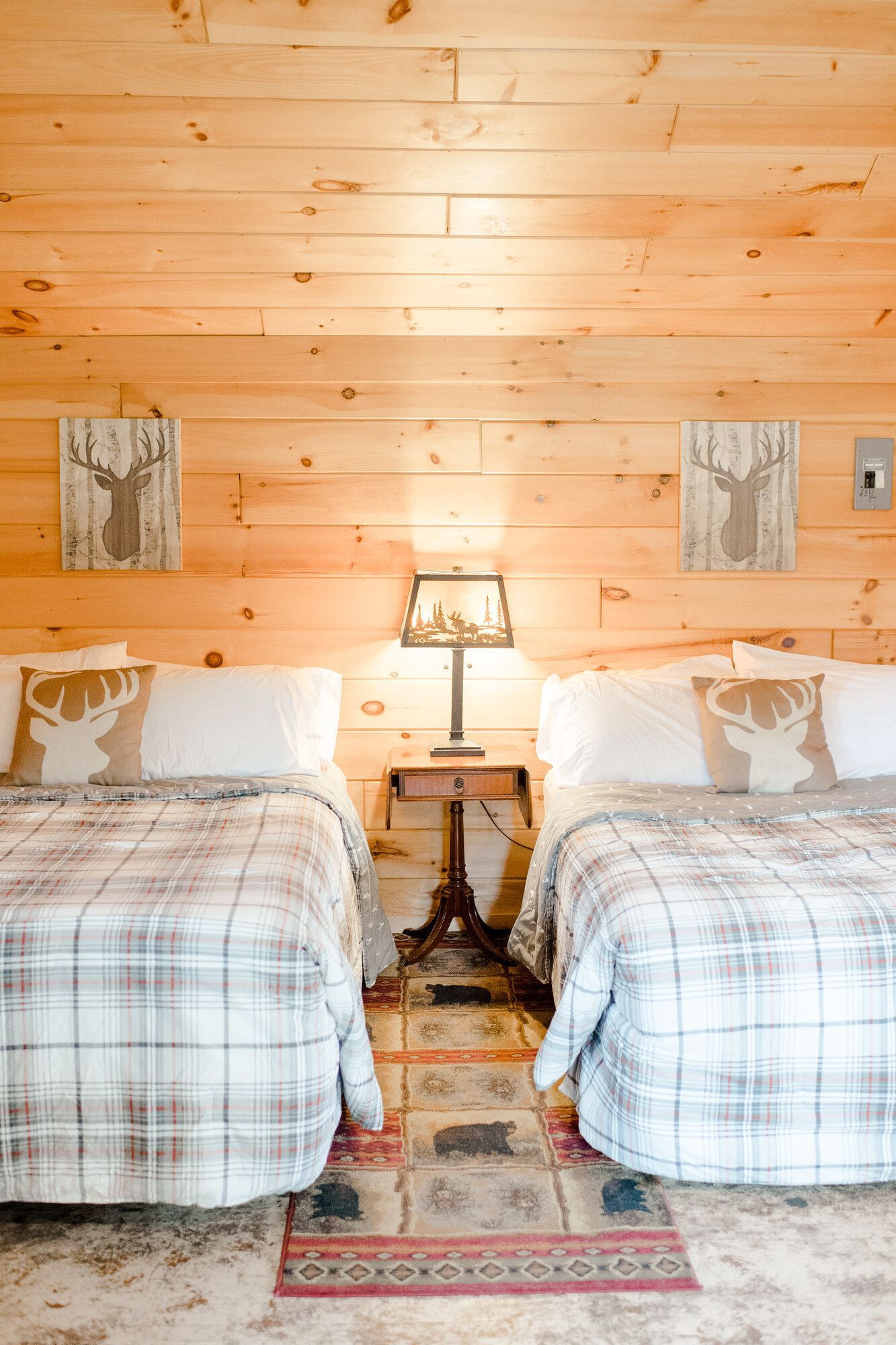 Cozy bedroom with two twin beds, plaid blankets, bedside lamp, and wood-paneled walls
