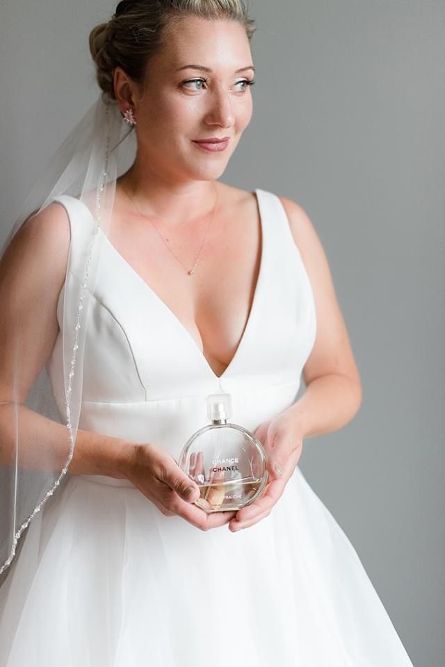 Bride in white gown holding a perfume bottle, standing against a gray background.