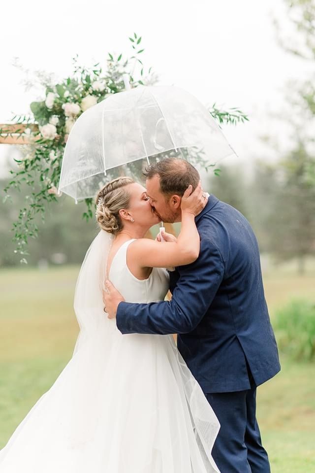 Couple kissing under a clear umbrella in a green outdoor setting, bride in white and groom in blue suit