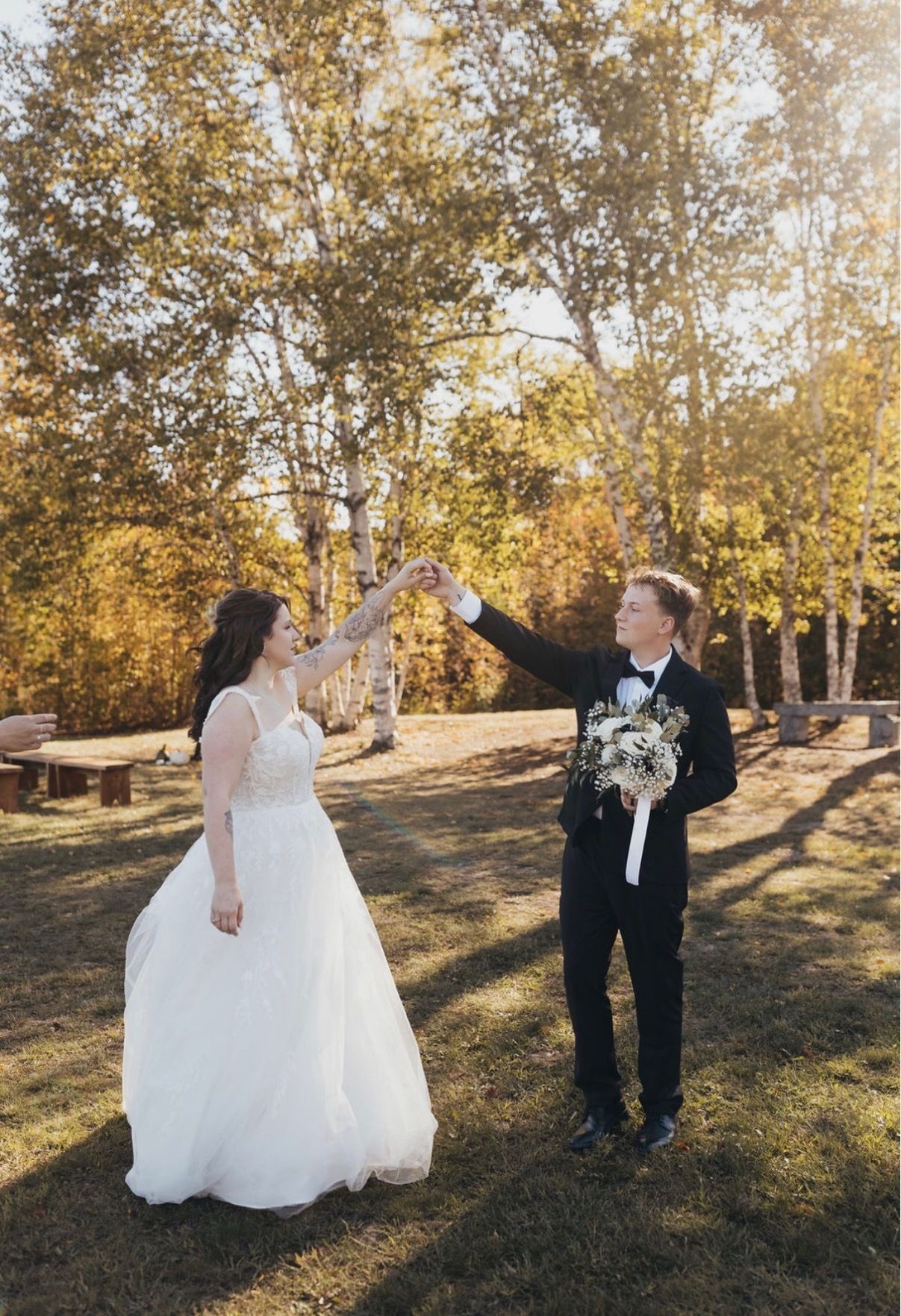 Bride and groom in white and black raising hands outdoors among autumn trees
