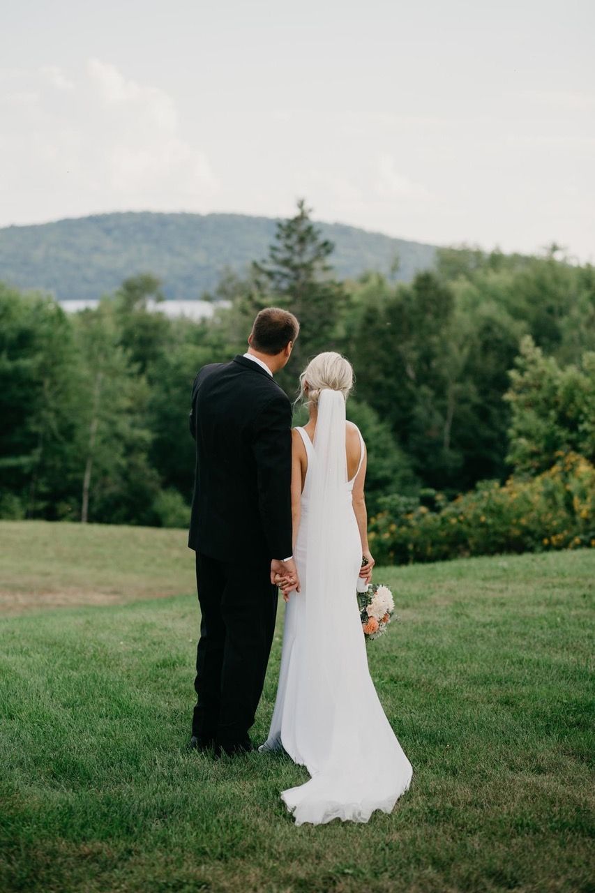 Bride and groom standing on a grassy hill, looking over a tree-lined landscape