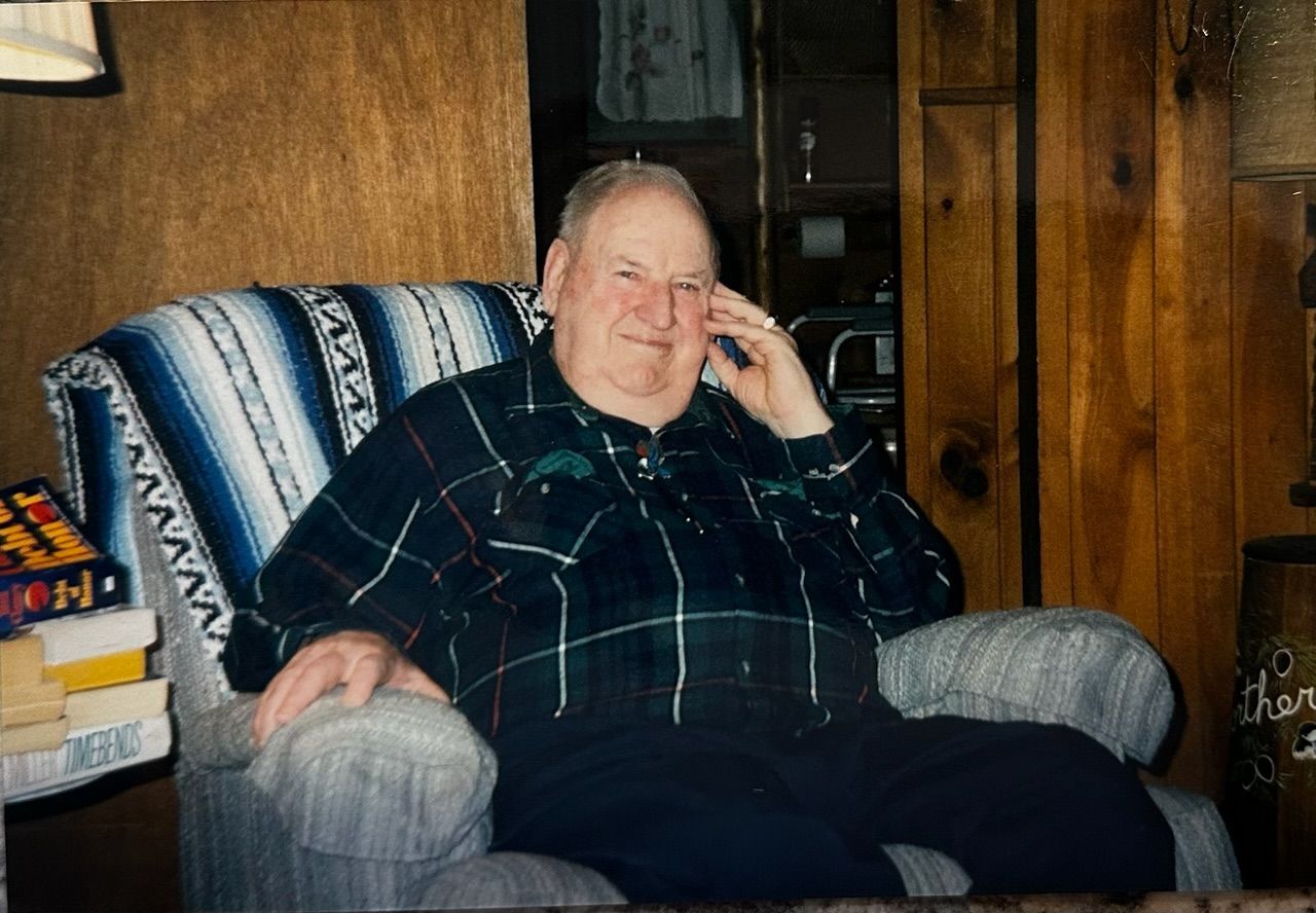 Older person sitting in a gray armchair, holding a phone to their ear in a wood-paneled room